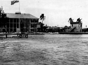 A three story building on a shoreline with a flagpole, trees and a dock out front.