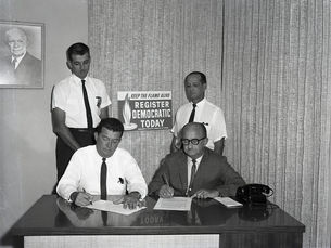 Two men sit at a desk writing with two more men standing behind them. A sign on the wall reads Keep the flame alive register Democratic today.