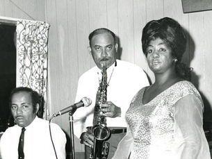 A man sitting at a piano, a man playing saxophone and a woman in front of a microphone.