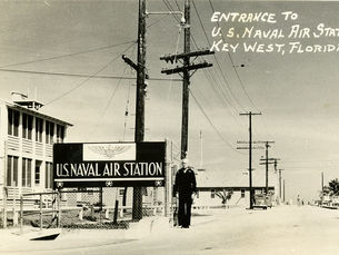 A sailor standing behind a sign that reads U.S. Naval Air Station. Writing on photo reads Entrance to U.S. Naval Air Station Key West, Florida.