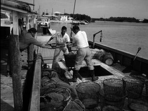 Two people unload a basket from a boat to another person on a dock.