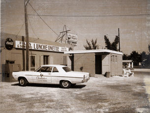 A 1960s style car in front of a building with a sign that reads Luncheonette