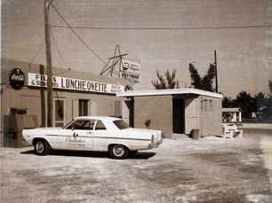 A 1960s style car in front of a building with a sign that reads Luncheonette