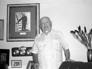 A man wearing a guayabera standing in a room with a photo of harry truman on the wall behind him.