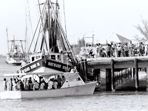 A small commercial fishing boat with a lot of people on it appraoches a dock that has a lot of people on it.