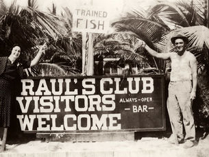 A man and woman standing in front of palm trees and a sign that reads Trained Fish. Raul's Club Visitors Welcome always-open bar.