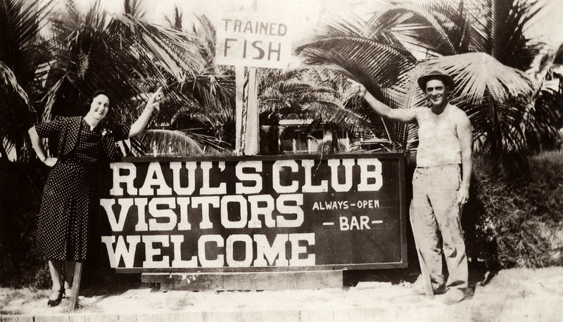 A man and woman standing in front of palm trees and a sign that reads Trained Fish. Raul's Club Visitors Welcome always-open bar.
