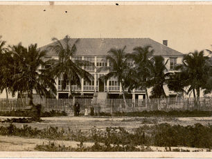 A large two story building with a fence, palm trees and several people in front of it.