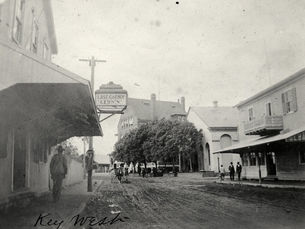 An unpaved street with people on the sidewalk and a sign that reads Last Chance Leon's on the left. Handwriting on the photo reads Key West.