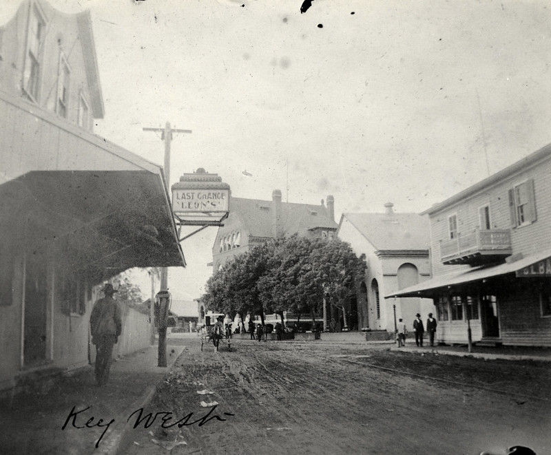 An unpaved street with people on the sidewalk and a sign that reads Last Chance Leon's on the left. Handwriting on the photo reads Key West.