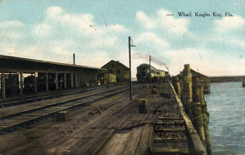 A postcard of a train approaching a station on the water. Text on the card reads Wharf, Knights Key Fla.