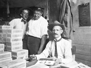 Two men standing and one sitting behind a table with cigar boxes piled on it.