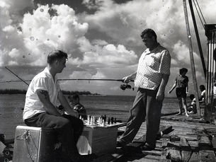 two men on a dock with fishing poles and a chess board.