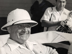 A man wearing a pith helmet smiles while sitting in the back of an open car
