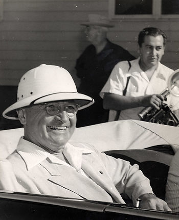 A man in a pith helmet smiles while sitting in the back of an open car
