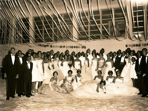 A group of elegantly dressed people in tuxedos and gowns pose in a decorated hall with streamers. 