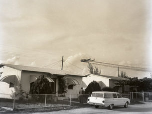 Houses with a station wagon parked out front and plane overhead trailing spray.