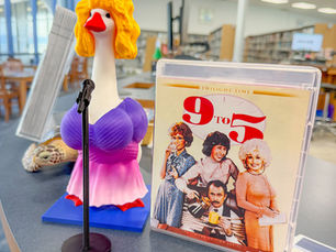 a goose figurine wearing a blond wig and purple an dpink dress next to the DVD for the movie 9 to 5.
