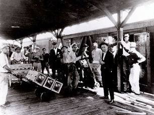 A group of people, two with boxes on dollies, on a platform next to a train car.