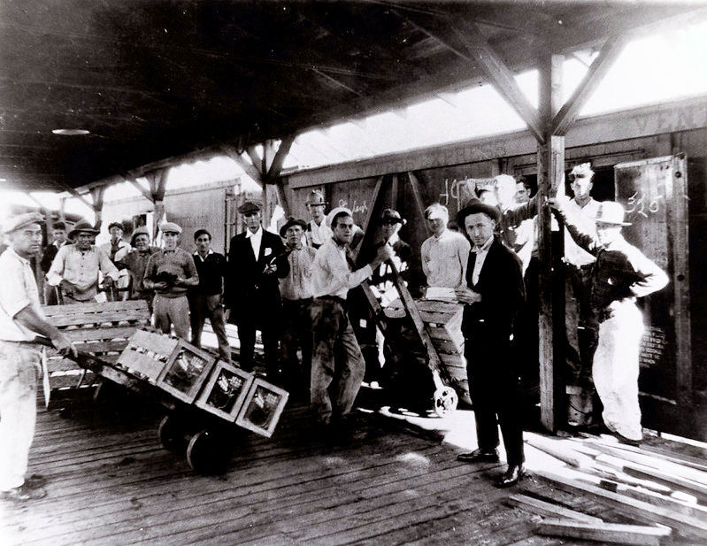A group of people, two with boxes on dollies, on a platform next to a train car.