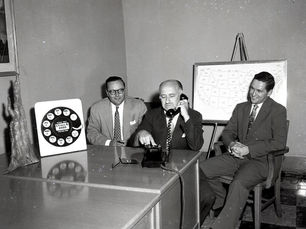 Three men sit behind a table while one dials a phone