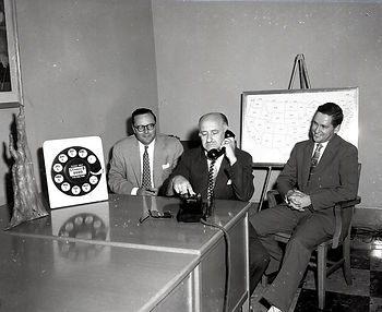 Three men sit behind a table while one makes a phone call.