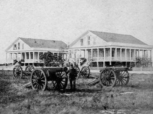 A man in uniform with several cannons mounted on wheels and two buildings in the background.