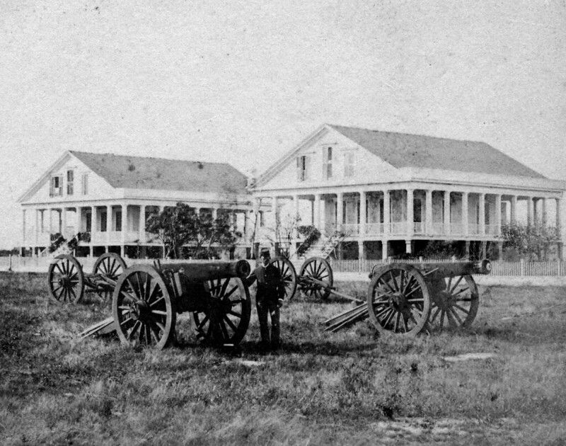 A man in uniform with several cannons mounted on wheels and two buildings in the background.