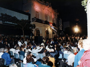 a crowd of people outside a building with a cuban flag flying from the balcony