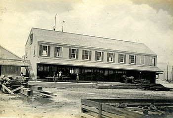 A two story building with an open area on the ground floor and several people and horses and carriages out front.