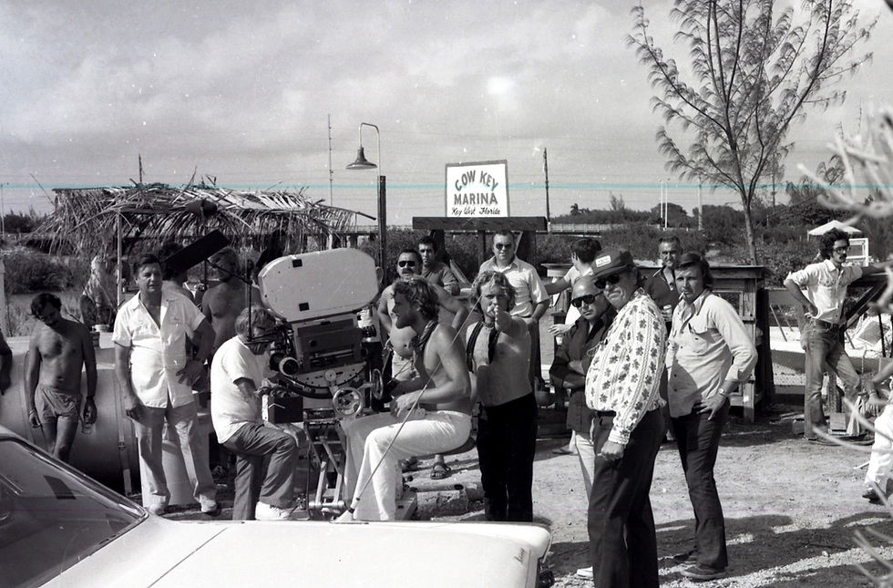 A group of people stand around a movie camera with a sign in the background that reads Cow Key Marina.